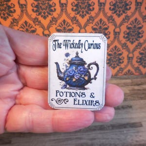 May include: A rectangular card with the text "The Wickedly Curious" and "Potions & Elixirs". The card features a blue teapot with purple roses. The background is a patterned orange and black design.