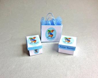 Könnte beinhalten: Ein Set aus zwei kleinen quadratischen weißen Geschenkboxen und einer weißen Geschenktüte mit blauen Henkeln, die alle mit einer Peter-Hase-Illustration versehen sind. Die Boxen haben einen blau karierten Rand und die Tüte enthält blaues Seidenpapier.
