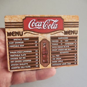May include: A vintage-style Coca-Cola menu card with a wooden background. The menu lists specials of the day, sandwiches, and desserts with prices. The menu includes items like clam chowder, sirloin steak, chicken pot pie, and chocolate layer cake.