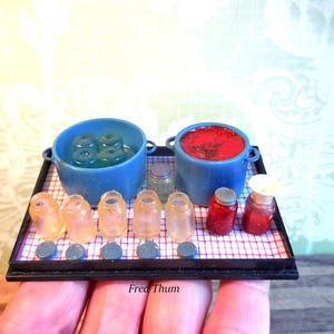 Op de afbeelding: Een zwart miniatuurdienblad met een rood-wit geblokt oppervlak met twee blauwe potten, vijf kleine doorschijnende potjes, twee rode potjes en vijf kleine blauwe ronde schijfjes. Eén blauwe pot bevat vier groene voorwerpen, de andere rode vloeistof.