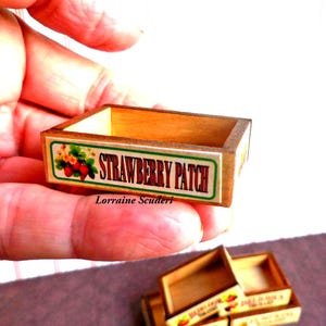 May include: A miniature rectangular wooden crate with a light brown finish. The front panel features a white label with a green border, red strawberry illustrations, and the black text STRAWBERRY PATCH. The name Lorraine Scuderi is printed below.