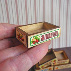 May include: A miniature wooden crate with a printed label that says "Strawberry Patch". The crate is brown with a white and green label. The label has a picture of strawberries and flowers.