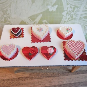 Può includere: Un tavolo bianco con sei torte a forma di cuore decorate con glassa rosa e bianca. Le torte sono su basi a forma di cuore rosse.