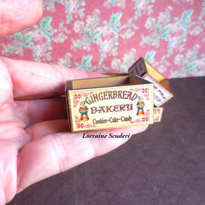 May include: A miniature wooden box with a gingerbread bakery sign. The sign features a red and white striped awning, gingerbread men, and the text "Gingerbread Bakery Cookies-Cake-Candy".