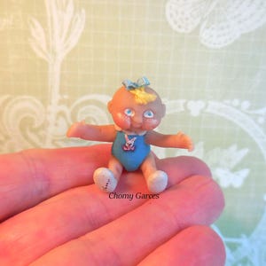 May include: A miniature doll with a light skin tone, blue eyes, and a small blue bow. The doll wears a blue onesie with a pink bunny graphic. The doll's arms are outstretched, and it has white shoes. The doll is sitting on a hand.