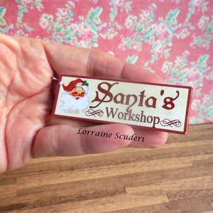 May include: A small wooden sign with a red background and white lettering that reads "Santa's Workshop". A cartoon image of Santa Claus is in the top left corner of the sign. The sign is held in a hand.