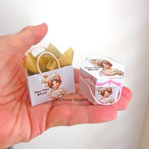 May include: A miniature white shopping bag and hexagonal box, both adorned with a vintage illustration of a woman in a hat. The bag contains gold tissue paper and has a pink handle. The box is tied with pink string and ribbon.