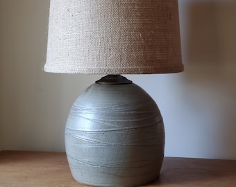 May include: A ceramic table lamp with a gray, textured base and a burlap lampshade.