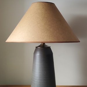 May include: A ceramic table lamp with a dark gray base and a beige lampshade. The lampshade is conical and has a brown trim. The lamp sits on a wooden surface. The lamp is designed for indoor use.