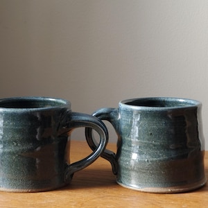 May include: Two dark green ceramic mugs with a glazed finish. The mugs have a rounded shape and a handle on each side.