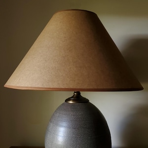 May include: A table lamp featuring a rounded, speckled gray ceramic base with a smooth finish and a metallic bronze-toned neck. The lamp is topped with a conical, tan-colored lampshade that has a simple, clean edge and a solid, neutral appearance.