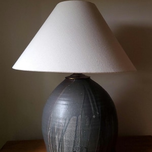 May include: A ceramic table lamp with a white lampshade. The base is a dark gray with a dripped glaze effect. The lamp has a brass-colored ring at the top of the base.