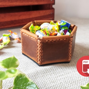 May include: A hexagonal leather trinket dish with a brown leather exterior and decorative stitching. The dish is filled with colorful candies.