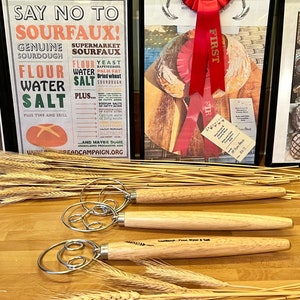May include: A display of baking-related items. A framed poster states "Say No To Sourfaux!" and lists ingredients. A framed photo shows a loaf of bread with a red "First" ribbon. Three dough whisks with wooden handles are in the foreground.