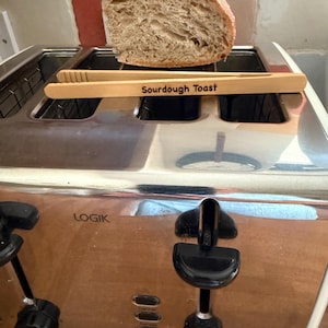 May include: A stainless steel LOGIK toaster with a slice of sourdough bread. Wooden tongs with the words "Sourdough Toast" are resting on the toaster. The bread is light brown with a textured surface.