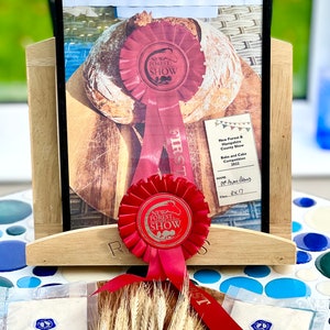May include: A display featuring a loaf of organic einkorn sourdough with malted wheat flakes, showcased with a red ribbon from the New Forest Show. The display includes packets of ingredients and wheat stalks, with text indicating a baking competition.
