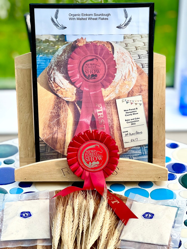 May include: A display featuring a loaf of organic einkorn sourdough with malted wheat flakes, showcased with a red ribbon from the New Forest Show. The display includes packets of ingredients and wheat stalks, with text indicating a baking competition.