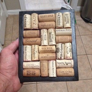 May include: A black framed cork board with a variety of wine corks arranged in a grid pattern. The corks are all different sizes and shapes, and some have labels with the names of wineries or vineyards. The board is a great way to display your love of wine.
