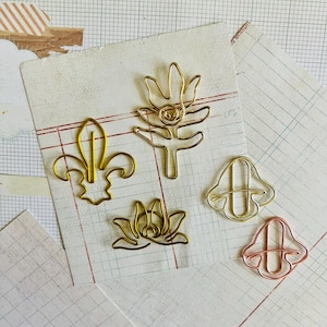 May include: Five gold-toned metal paper clips shaped like different flowers and plants. The clips are on a white sheet of paper with a grid pattern.