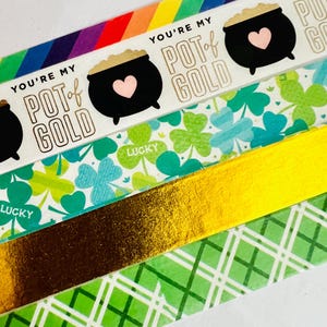 May include: Four rolls of washi tape with a St. Patrick's Day theme. The top roll has a rainbow background with the text "You're my pot of gold" and a black pot of gold with a pink heart. The second roll has a white background with green shamrocks and the text "Lucky". The third roll is gold foil. The fourth roll has a green and white checkered pattern.