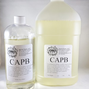 May include: Two clear plastic bottles of CAPB liquid surfactant from Spiral Eyes Soap. The smaller bottle contains 32oz/908ml, and the larger bottle is a gallon size. Both bottles have white labels with product information.