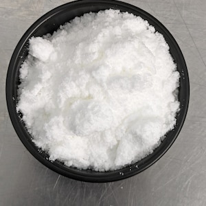 May include: A top-down view of a black bowl filled with a large quantity of white, powdery substance. The substance has a fluffy, cloud-like appearance, with a slightly uneven surface. The bowl is round and has a matte finish.