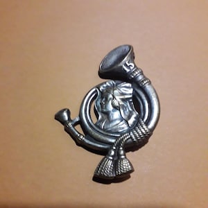 May include: A silver-toned metal badge with a stylized image of a woman's head and a horn. The number "15" is visible on the horn.