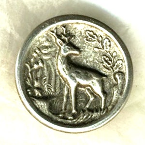 May include: A silver-toned button with a raised design of a deer standing in a field of grass with a tree branch behind it.