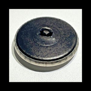May include: The back of a silver button with a loop on top. The button has a textured edge and a smooth, dark gray center.