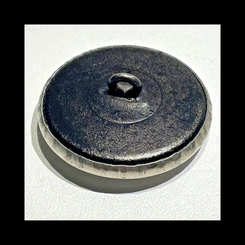 May include: The back of a silver button with a loop on top. The button has a textured edge and a smooth, dark gray center.