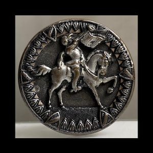 May include: A round, metallic button featuring a raised relief design of a figure on horseback holding a flag. The central motif is surrounded by a decorative border of repeating triangular shapes. The finish is dark silver with highlights.
