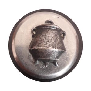 May include: A silver-colored, circular button with a raised image of a cauldron. The cauldron has a lid and two handles. The button has a dark, circular border.