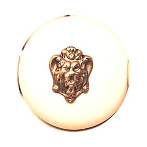 May include: A round, off-white cabinet knob with a bronze-colored lion's head emblem in the center. The lion's head has detailed features and is slightly raised from the surface of the knob. The knob has a smooth, polished finish.