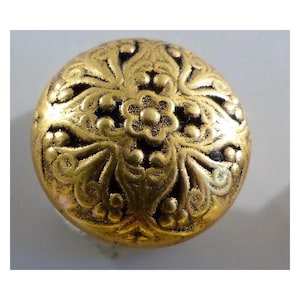 May include: A round, antique-style button with a gold finish. The button features an embossed floral design with a central flower and surrounding leaves and swirls. The button's surface has a slightly aged appearance.