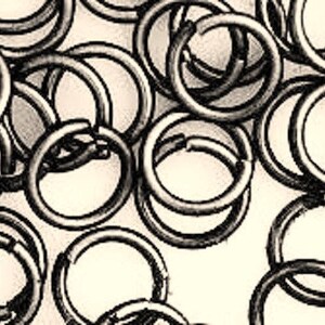 May include: Close-up of a collection of silver-colored jump rings, each with a circular shape and a small opening. The rings are scattered across a light beige background, creating a textured pattern. Ideal for jewelry making and crafting.