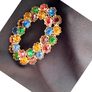 May include: A gold-toned brooch with a circular design featuring a variety of colourful rhinestones. The rhinestones are arranged in a circular pattern and are set in a gold-toned metal.