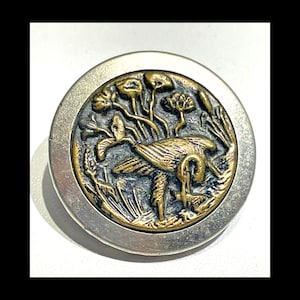 May include: A silver and gold button with a raised design of a bird in a marsh. The bird is in the center of the button and is surrounded by reeds and flowers.