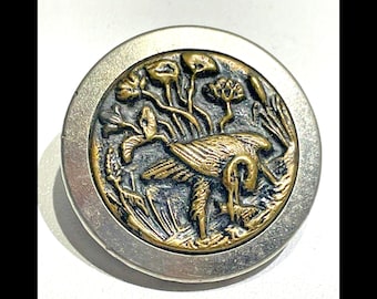 May include: A silver and gold button with a raised design of a bird in a marsh. The bird is in the center of the button and is surrounded by reeds and flowers.