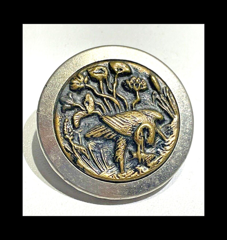 May include: A silver and gold button with a raised design of a bird in a marsh. The bird is in the center of the button and is surrounded by reeds and flowers.