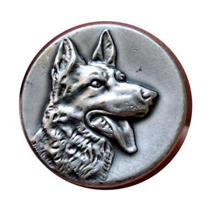 May include: A round, silver-toned metal button featuring a detailed relief of a German Shepherd dog's head. The dog is depicted with its mouth open and tongue out, set against a plain background. The button has a slightly aged appearance.