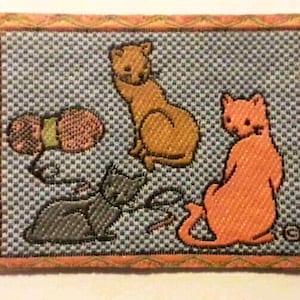 Può includere: Un toppa di tessuto blu e bianco con motivo a righe con tre gatti ricamati. Un gatto è arancione, uno è marrone e uno è grigio. I gatti sono tutti in pose diverse. La toppa ha un bordo rosa.