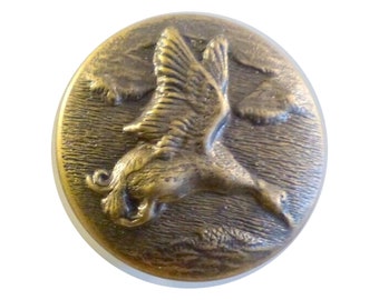 May include: A round, metallic button with a brass-toned finish featuring a raised, embossed design of a bird in flight. The background of the button has a textured, horizontal lined pattern that mimics water or sky, creating a relief effect.