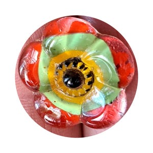 Peut inclure: Une fleur en verre colorée avec des accents rouges, jaunes, verts et noirs. La fleur a un centre circulaire avec un œil noir et des pétales jaunes.