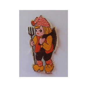 Könnte beinhalten: Gestickter Aufnäher mit einem Cartoon-Charakter, der einen rot-weiß gestreiften Hut, ein orangefarbenes Hemd, eine braune Hose und orangefarbene Schuhe trägt. Die Person hält eine braune Heugabel.
