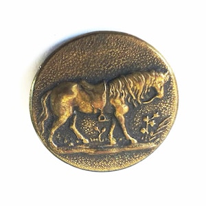 May include: A round, antique brass button with a detailed relief of a horse. The horse is saddled and walking, with floral accents in the background. The button has a textured surface and a vintage aesthetic.