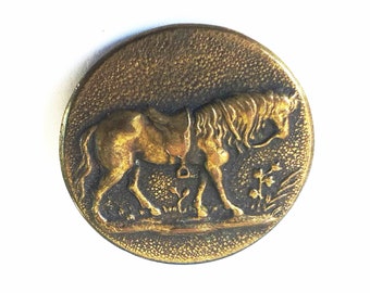 May include: A round, antique brass button with a detailed relief of a horse. The horse is saddled and walking, with floral accents in the background. The button has a textured surface and a vintage aesthetic.