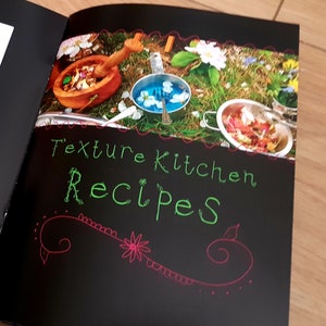 Puede incluir: Un libro negro con el t&iacute;tulo "Texture Kitchen Recipes" escrito en verde y rosa en la portada. La portada presenta una foto de un cuenco de madera, cuencos de metal y flores sobre un lecho de hierba verde.