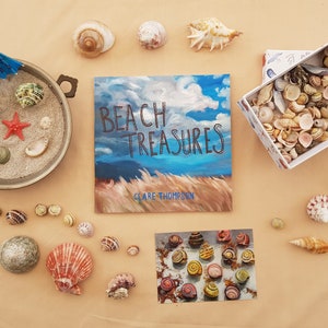 May include: A flat lay of seashells, a book titled "Beach Treasures" by Clare Thompson, and a small box filled with seashells. The book cover features a painting of a blue sky with white clouds and tall grass.