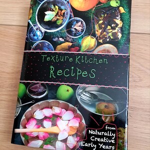 Puede incluir: Una portada de libro en blanco y negro con el t&iacute;tulo "Texture Kitchen Recipes" en letras verdes y rosas. La portada presenta una variedad de im&aacute;genes de comida y naturaleza, incluyendo manzanas, hojas y un taz&oacute;n de p&eacute;talos de rosa. El libro es de "Naturally Creative Early Years".