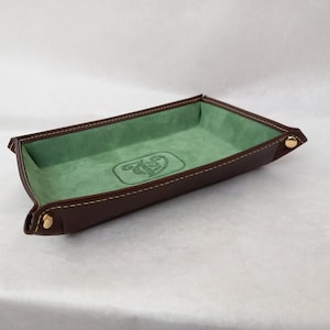 May include: A brown leather tray with a green felt lining and a gold button on each side. The tray has a green embroidered logo in the center.